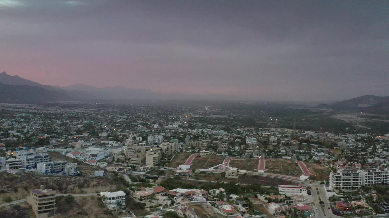 Los Cabos aerial view one second in, showing a spread of buildings and roads below a dark cloud ceiling, with mountain silhouettes and no visible people.