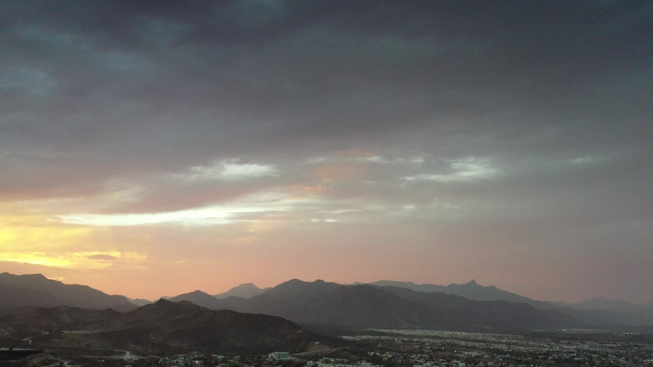 Los Cabos, Mexico sunrise view with dark mountain silhouettes, a low city grid, yellow light at the left horizon, and no visible people.
