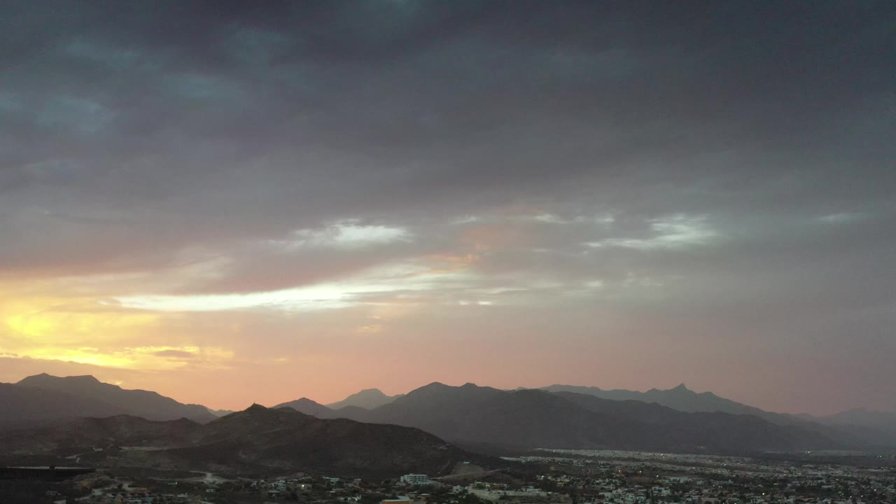 Los Cabos city blocks sit beneath layered mountain silhouettes, with orange light on the left horizon and a dense gray cloud ceiling, no visible people.
