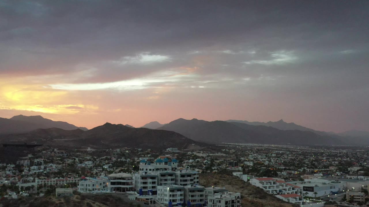 Los Cabos, Mexico appears from an elevated view with white and blue condominium buildings in front of the city grid and mountains under a dim sunset sky, with no visible people.