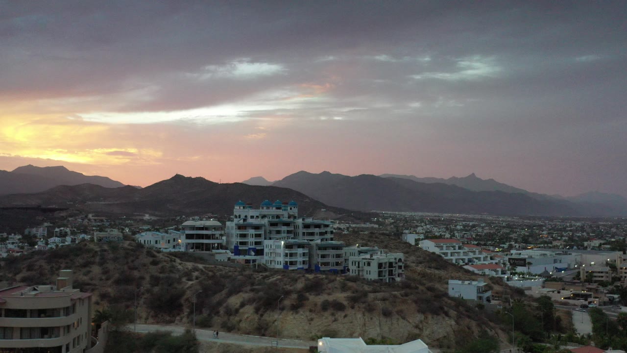 Wide view of Los Cabos, Mexico with hillside homes, white buildings, distant mountains, warm sunrise light at left, and no visible people.
