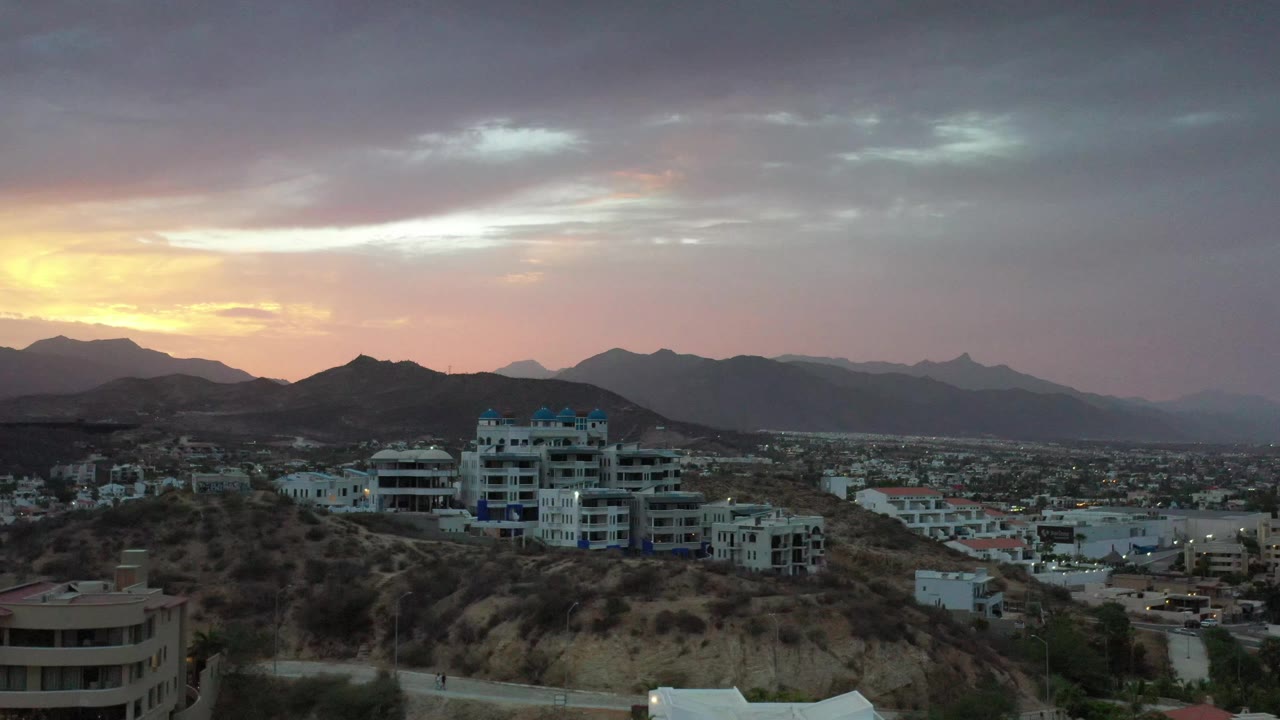Los Cabos hillside neighborhood seen from above, with a large white building near center, mountain ridges in the background, and no visible people.