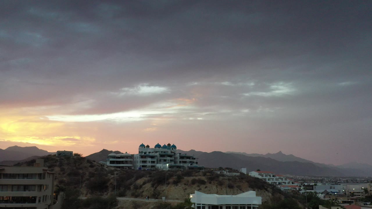 Los Cabos sunrise view with warm light at left, dark clouds overhead, hillside buildings below, distant mountains, and no visible people.