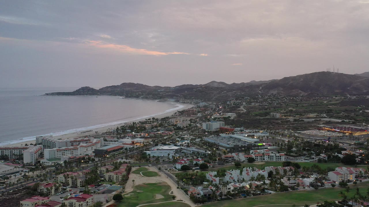 Wide Los Cabos, Mexico aerial frame with a curved beach and ocean on the left, town buildings and golf course below, hills in the distance, and no visible people.