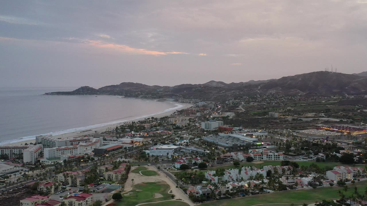 Los Cabos aerial view one second in, showing the pale surf line along the bay, clustered buildings near the shore, golf greens in the foreground, and no visible people.