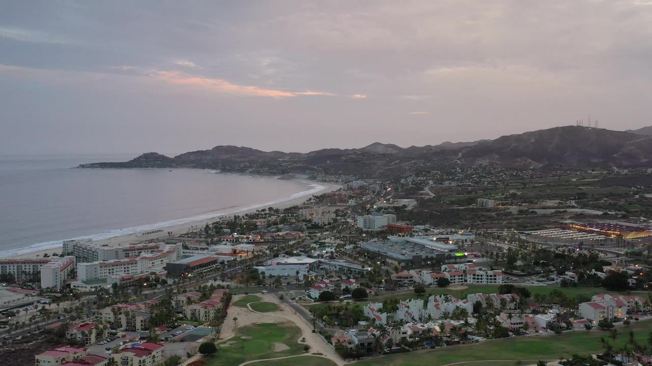 Aerial coastline view with beach arc, dense town area, hills, and muted sunset clouds.
