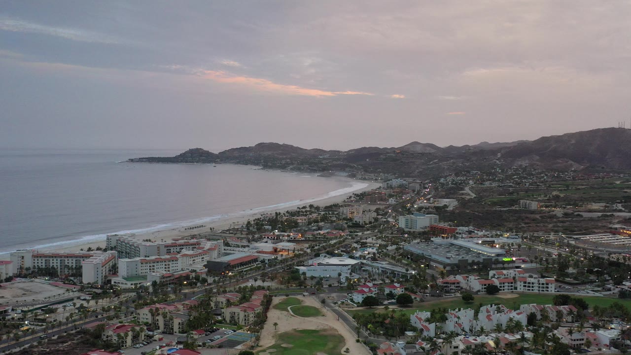 Wide aerial view of a coastal city beside a curving beach under cloudy evening sky.