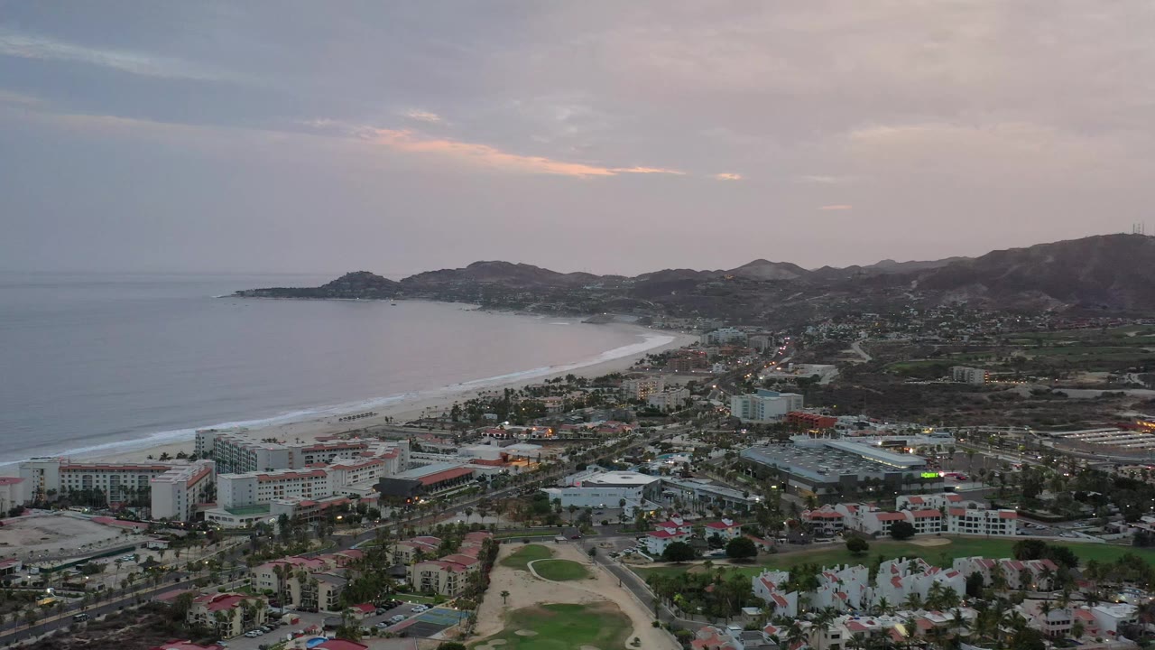 Aerial frame of Los Cabos, Mexico, showing the beach arc slightly farther across the center, ocean on the left, low buildings below, distant hills, evening clouds, and no visible people.
