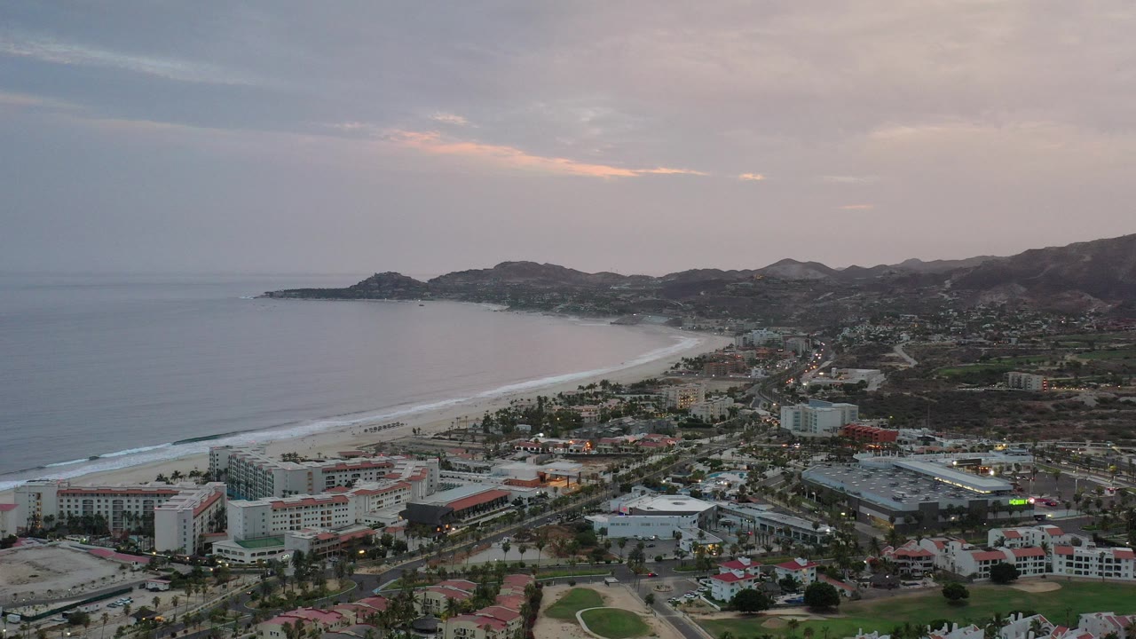Aerial view of coastline, beachfront buildings, roads, and hills under a cloudy sky.