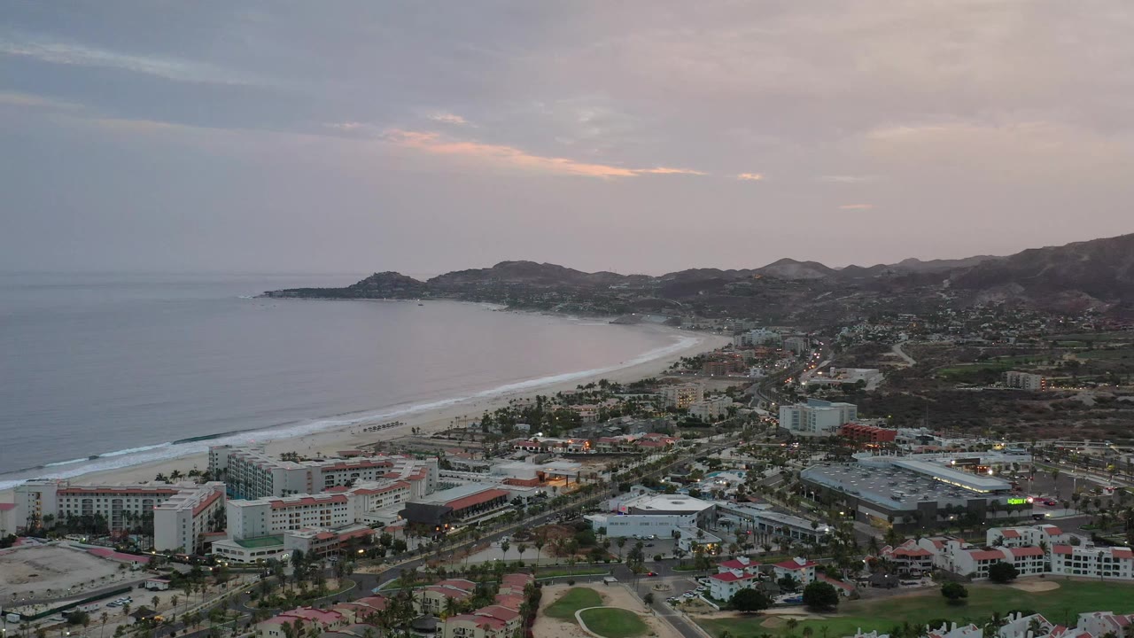 Aerial frame of Los Cabos, Mexico, showing more of the beachfront hotel area below the bay, with pale surf along the sand and no visible people.
