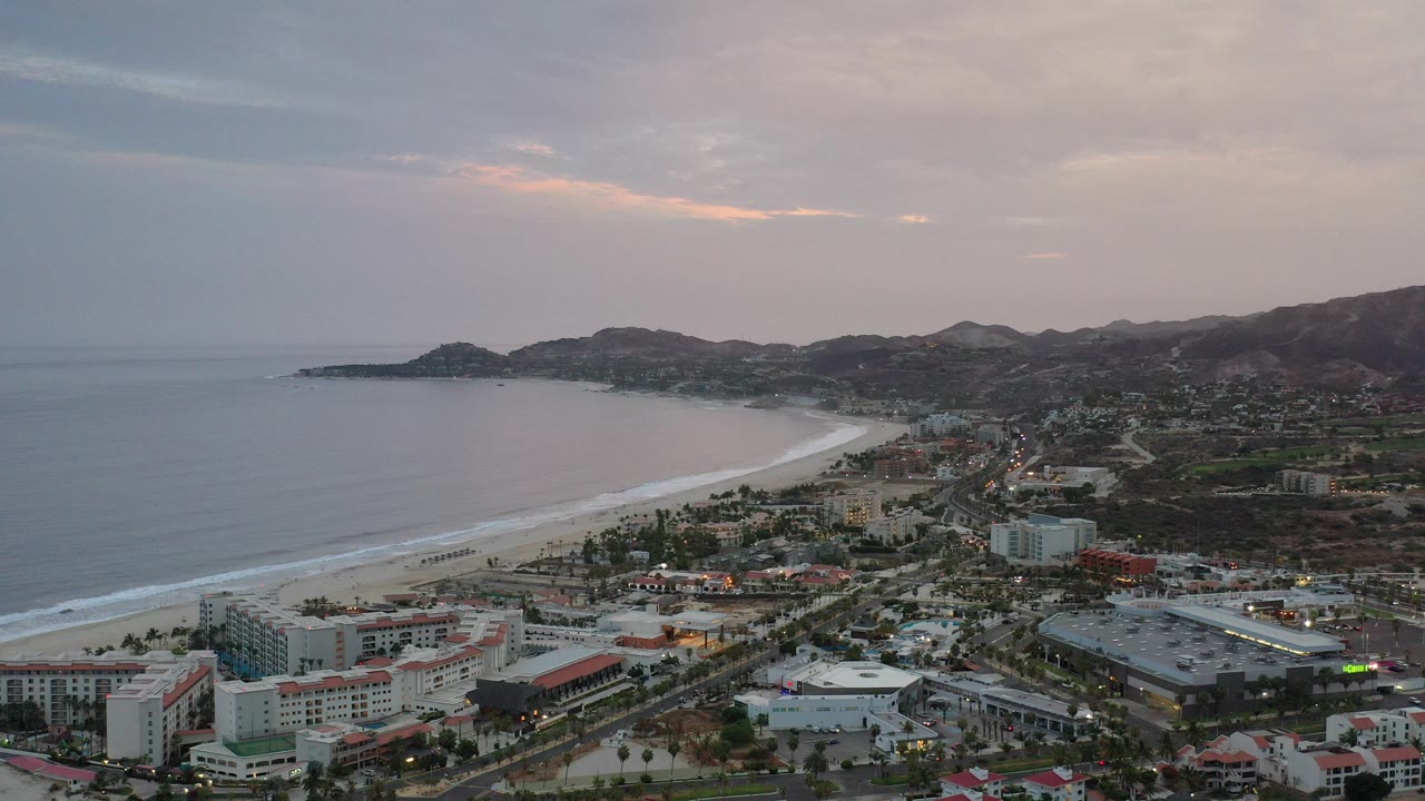 Aerial view of ocean, curved beach, city buildings, and hills at dusk.