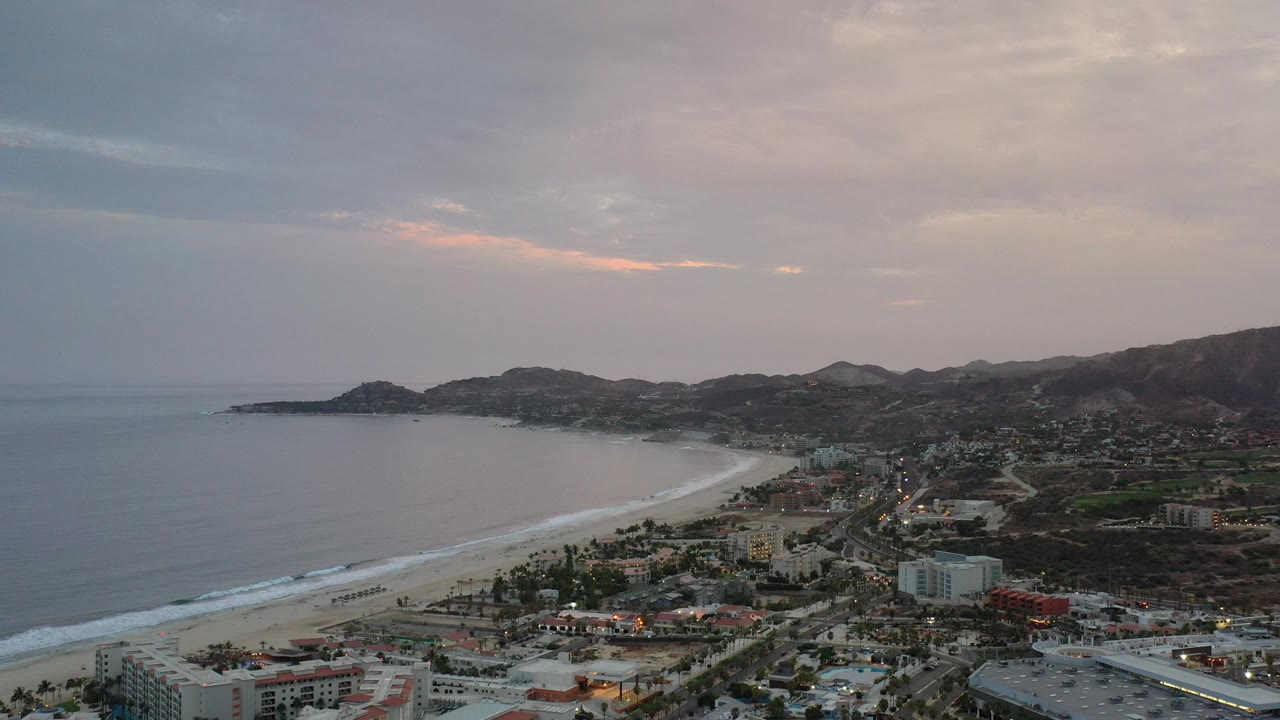 Aerial view of a curving beach, ocean, town, and hills under a gray evening sky.