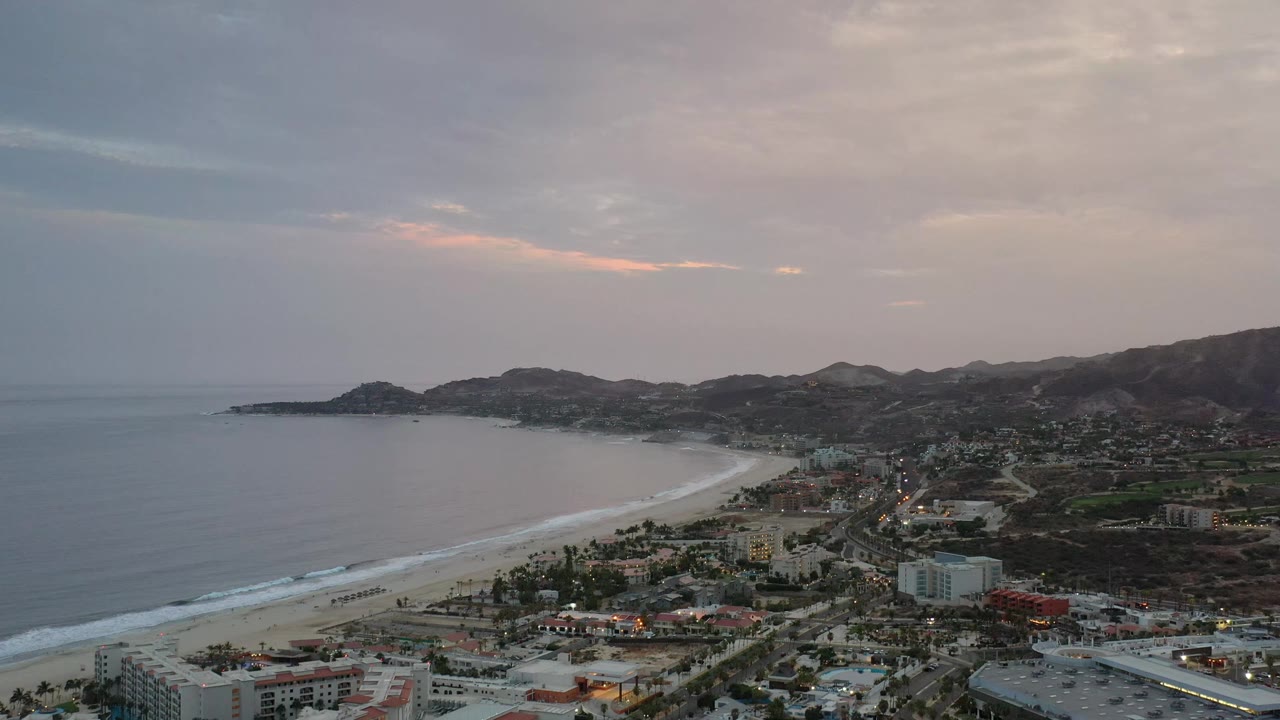 Aerial frame of Los Cabos, Mexico, looking along the shoreline as small surf bands meet the beach, with clustered buildings, inland roads, low hills, and no visible people.