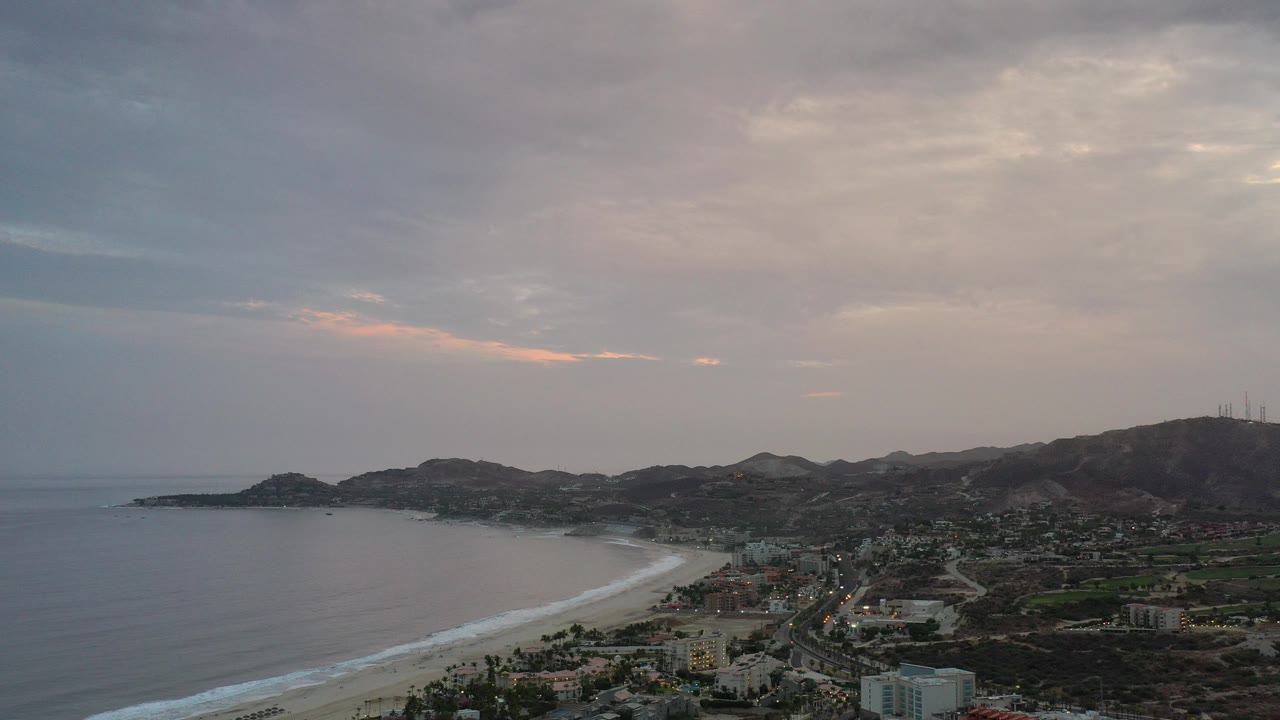 Aerial view of a curved beach, ocean, town buildings, hills, and cloudy sky.