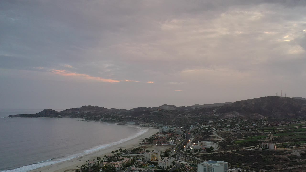 Wide aerial coastline with beach, town, hills, and cloudy evening sky.