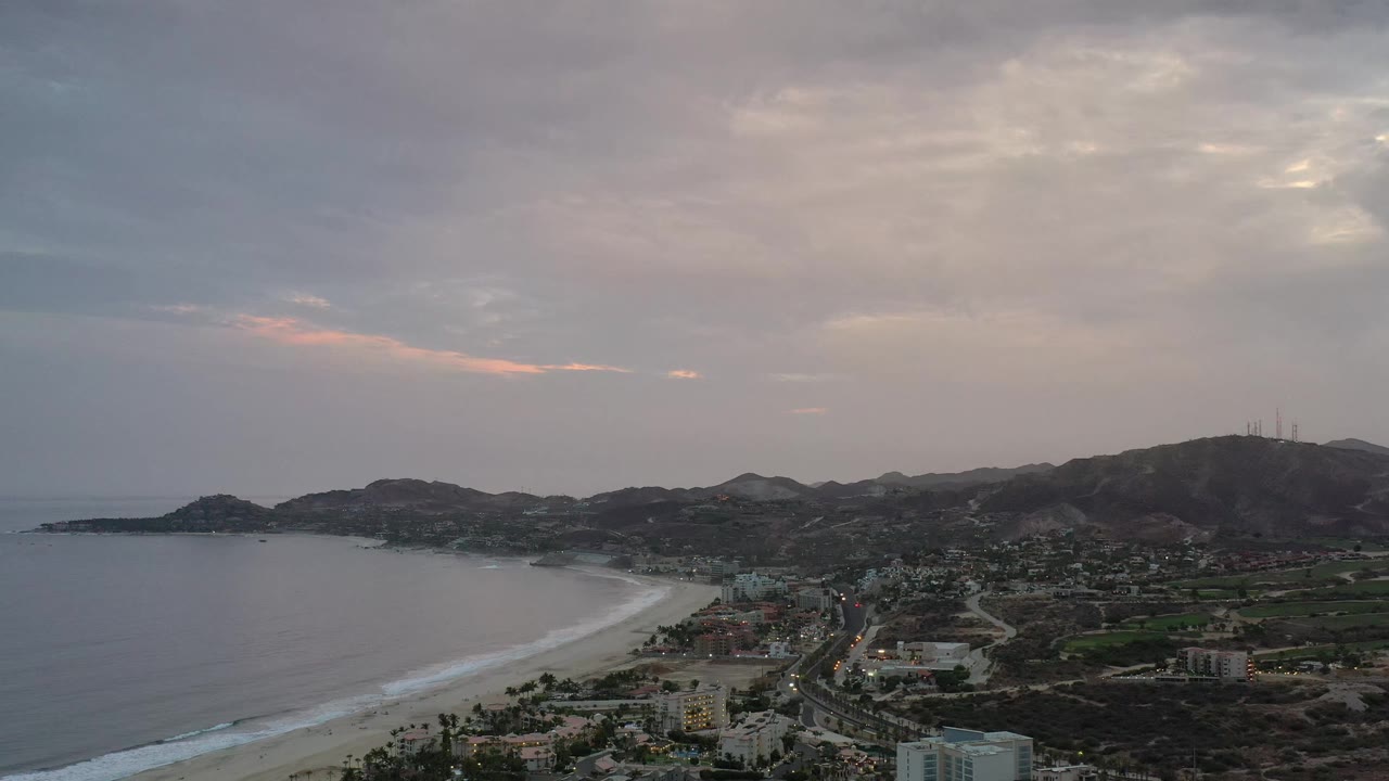 Elevated Los Cabos view with a curving beach at left, town streets below the hills, soft pink light in the clouds, and no visible people.