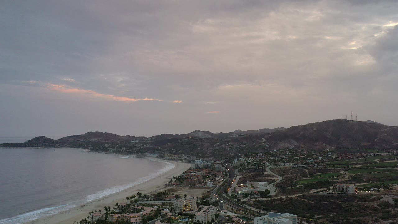Opening aerial frame over Los Cabos, Mexico, showing the curved beach and ocean at left, a coastal town and road below, dark mountains beyond, and no visible people.