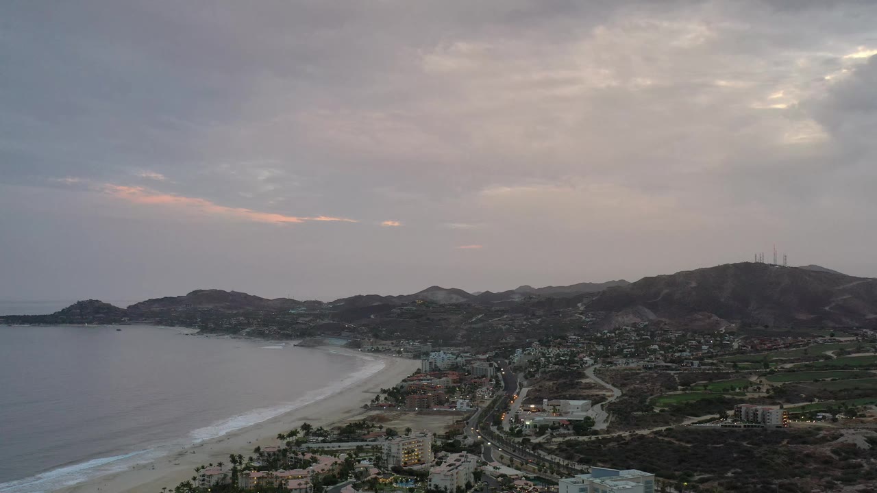 Aerial view over Los Cabos, Mexico, with the surf line stretching along the left edge, town blocks and a road in the center, hills behind, and no visible people.