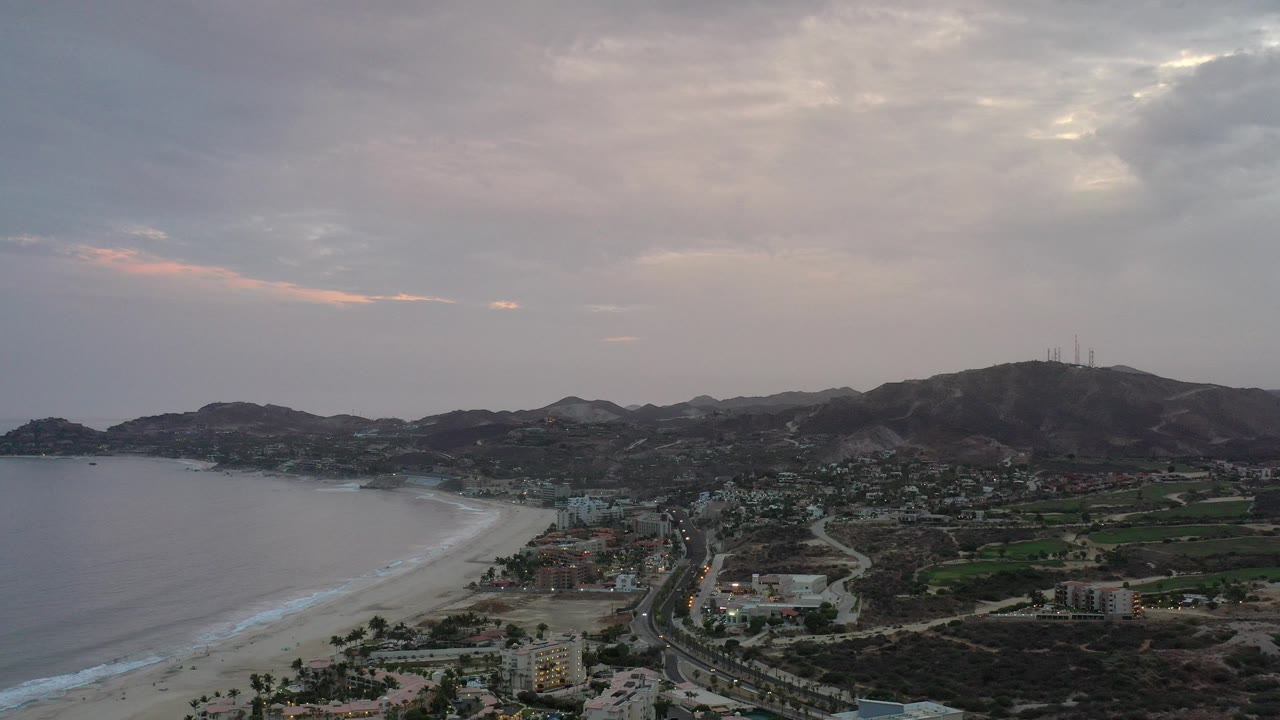 High aerial view over the Los Cabos coastline with ocean and surf at left, beach below, roads and buildings inland, hills on the horizon, and no visible people.