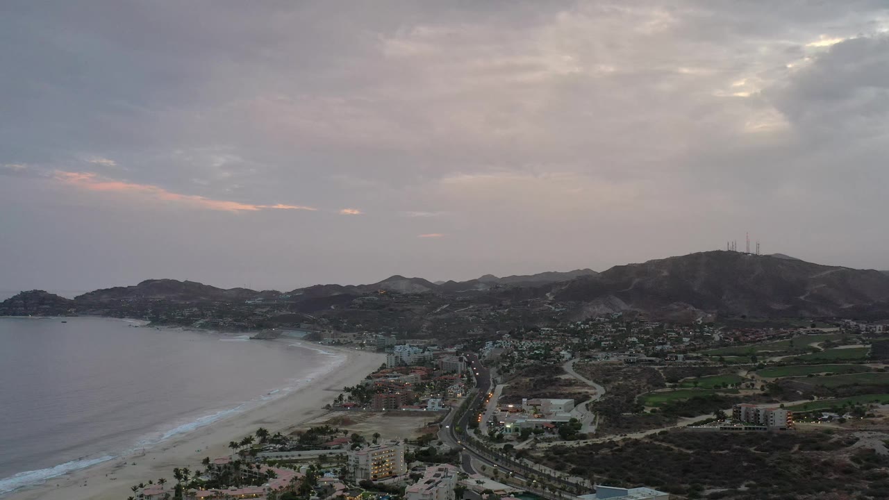 Aerial frame of Los Cabos with the beach curving along the left side, scattered buildings and roads through the center, green fields inland, a hill ridge beyond, and no visible people.