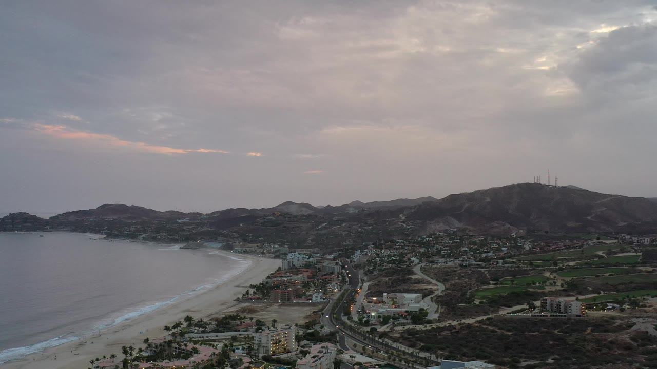 Coastal aerial with ocean, beach, town streets, green land, hills, and cloudy sunset light.