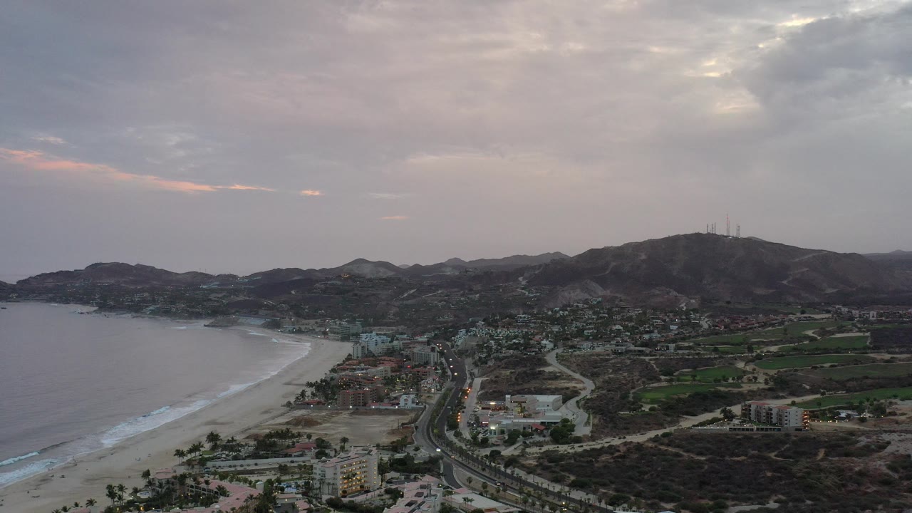 Wide aerial view of beach, coastal road, town buildings, and mountains under cloudy sky.
