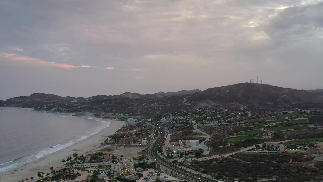Los Cabos coastline view one second in, showing the pale surf line along the beach, clustered buildings near the road, green parcels inland, dark hills, and no visible people.