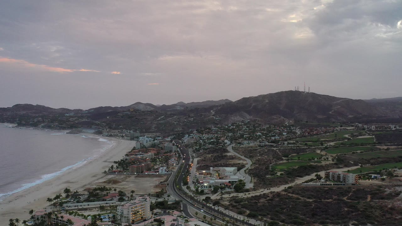 Opening aerial frame over Los Cabos, Mexico, showing ocean and a curving beach at left, road and buildings below, green fields and hills beyond, with no visible people.