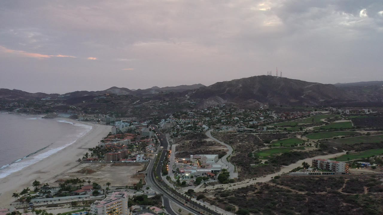 Aerial view of beach, coastal road, houses, fields, and hills under a cloudy sky.