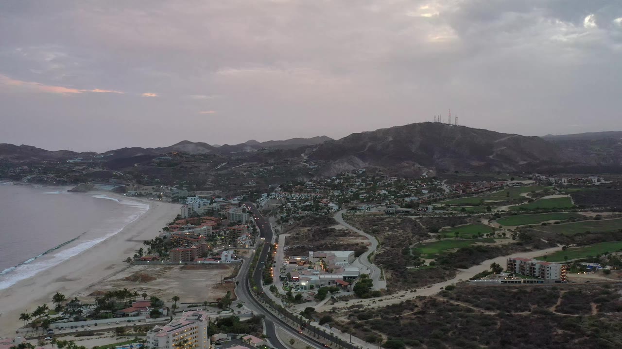 Aerial frame over Los Cabos, Mexico with the sandy beach and bay on the left, town blocks near the road, green fields to the right, and no visible people.