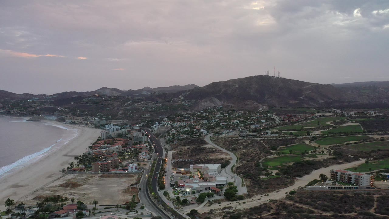 Wide aerial view of beach, road, buildings, fields, and hills under clouds.