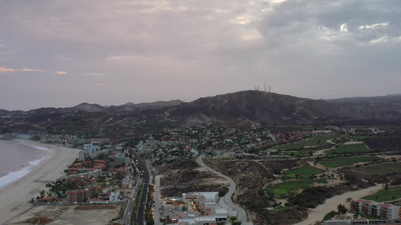 Opening aerial frame over Los Cabos, Mexico, with beach and surf at left, city blocks and roads below, hills with towers in the distance, and no visible people.