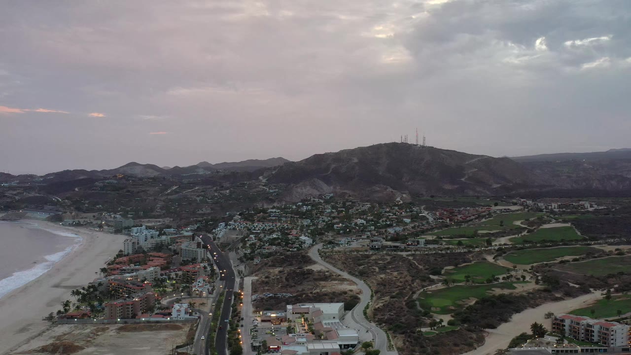 Aerial view of Los Cabos, Mexico, with the coastline still along the left edge, a main road and low buildings below, green patches inland, and no visible people.