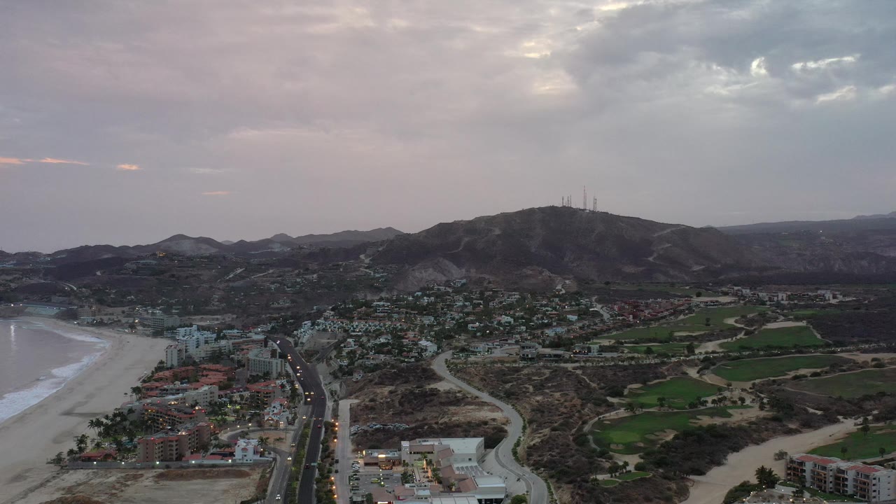 Coastal aerial with town, curved beach, green land, and mountain ridge.