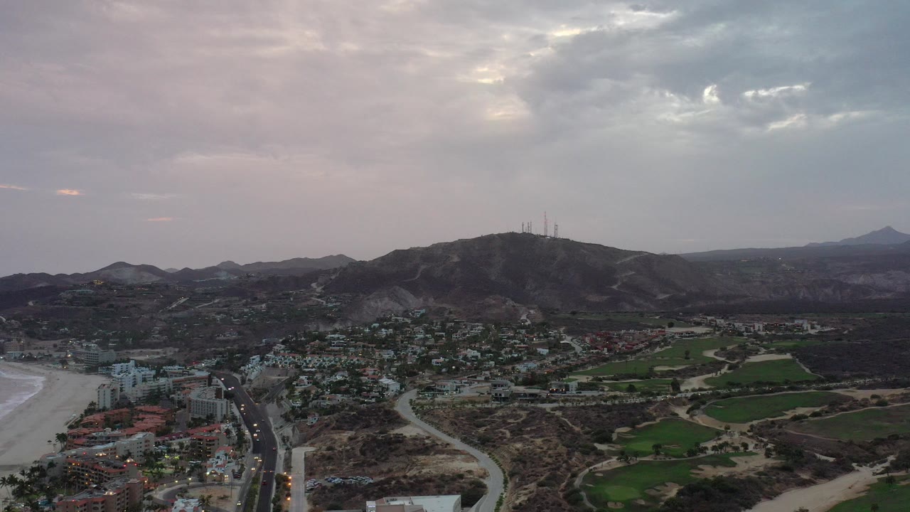 Aerial view over Los Cabos, Mexico, with buildings and roads below, a hillside with antenna structures near center, distant mountains, cloudy pink-gray sky, and no visible people.