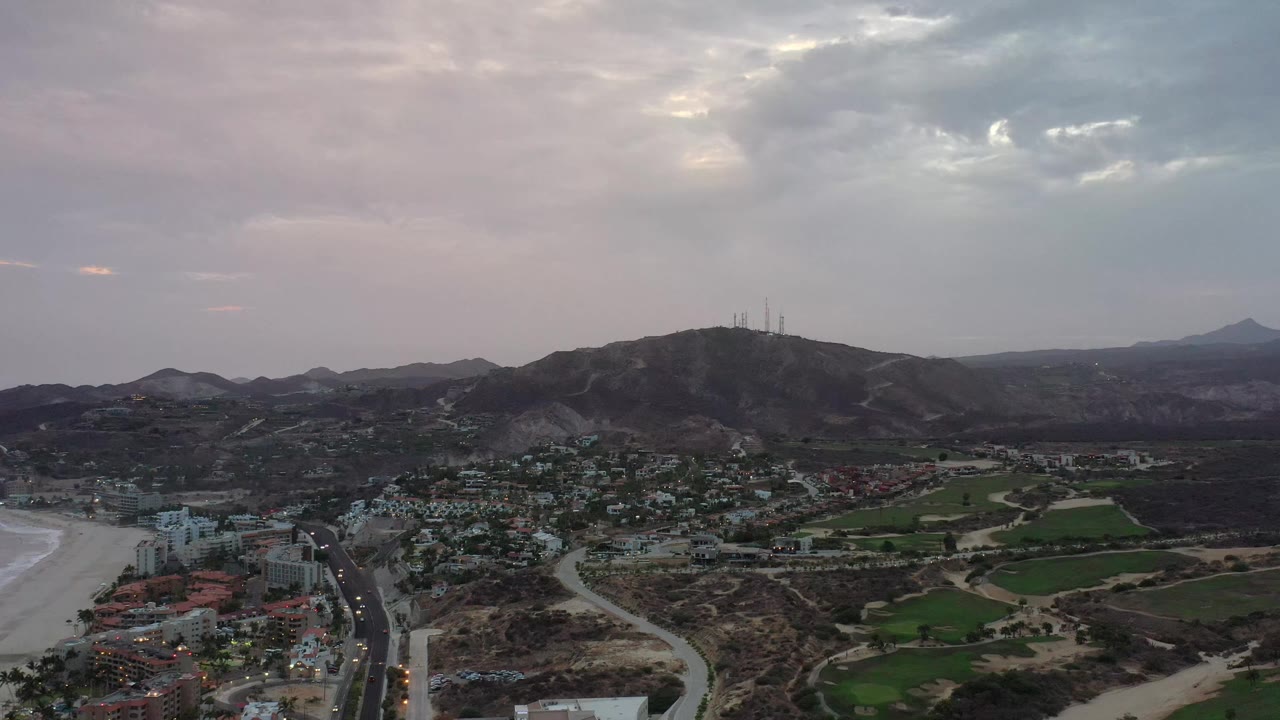 Wide Los Cabos aerial frame showing the road grid and clustered buildings at lower left, green fields in the valley, a dark hill with antennas, distant mountains, and no visible people.