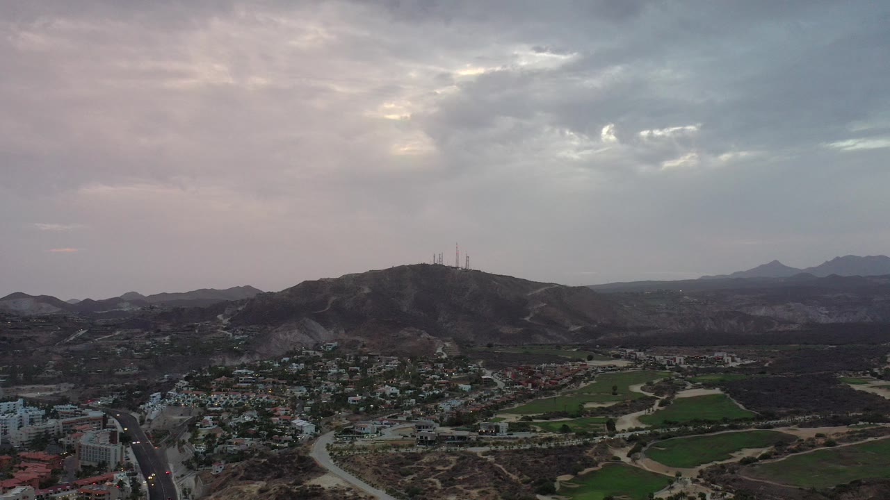 Wide aerial view of hills, buildings, roads, fields, and clouded sky.