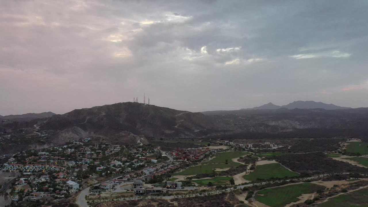 Los Cabos aerial view at 0:01 showing residential blocks in the lower frame, roads through dry terrain, green field patches, hills with towers, and no visible people.