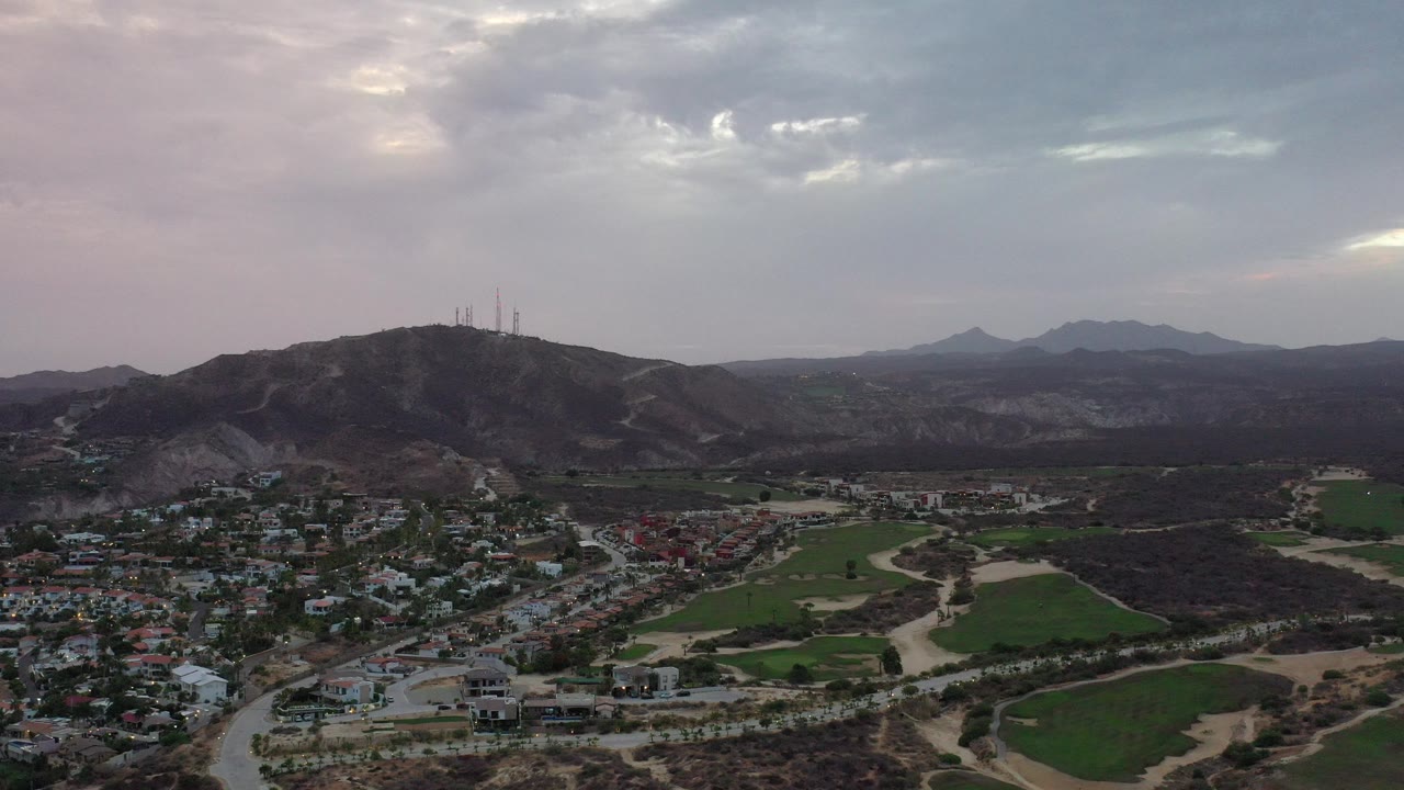 Aerial view of a hillside village, green fields, roads, and distant mountains under clouds.