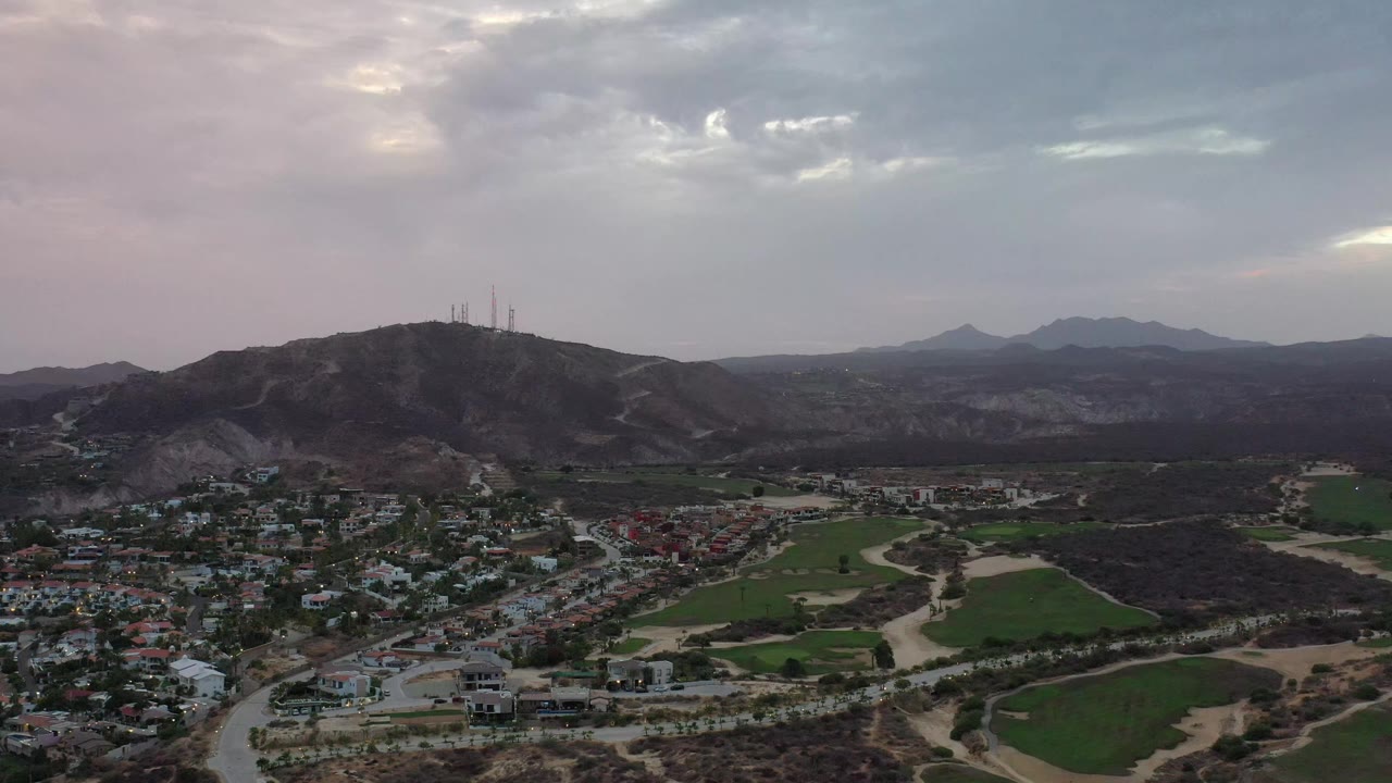 Aerial view in Los Cabos, Mexico, with clustered homes in the lower frame, green plots to the right, a tower-lined hill in the middle distance, and no visible people.