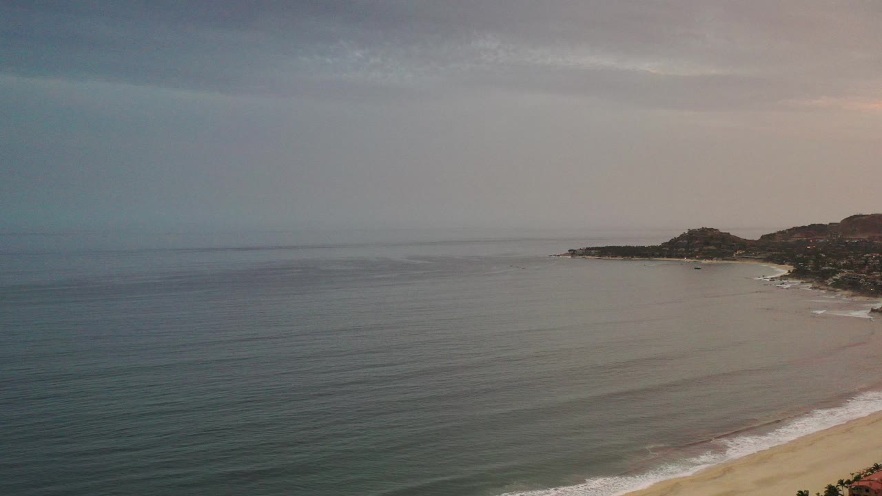 Wide Los Cabos coastal view over muted blue water, sandy shoreline at lower right, distant low hills, hazy sky, and no visible people.