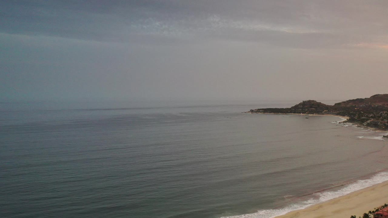Los Cabos ocean horizon with soft gray blue water, a strip of white surf along the beach, distant headland, warm haze, and no visible people.