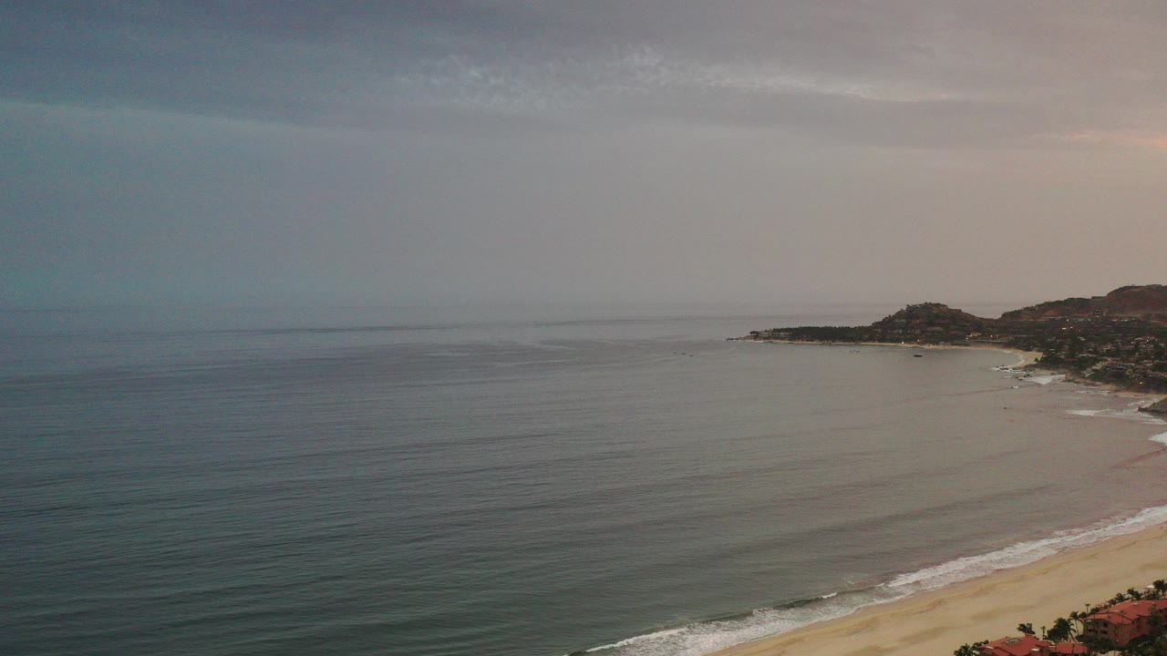 Wide aerial view of ocean, sandy beach, low hills, and red-roofed coastal buildings.