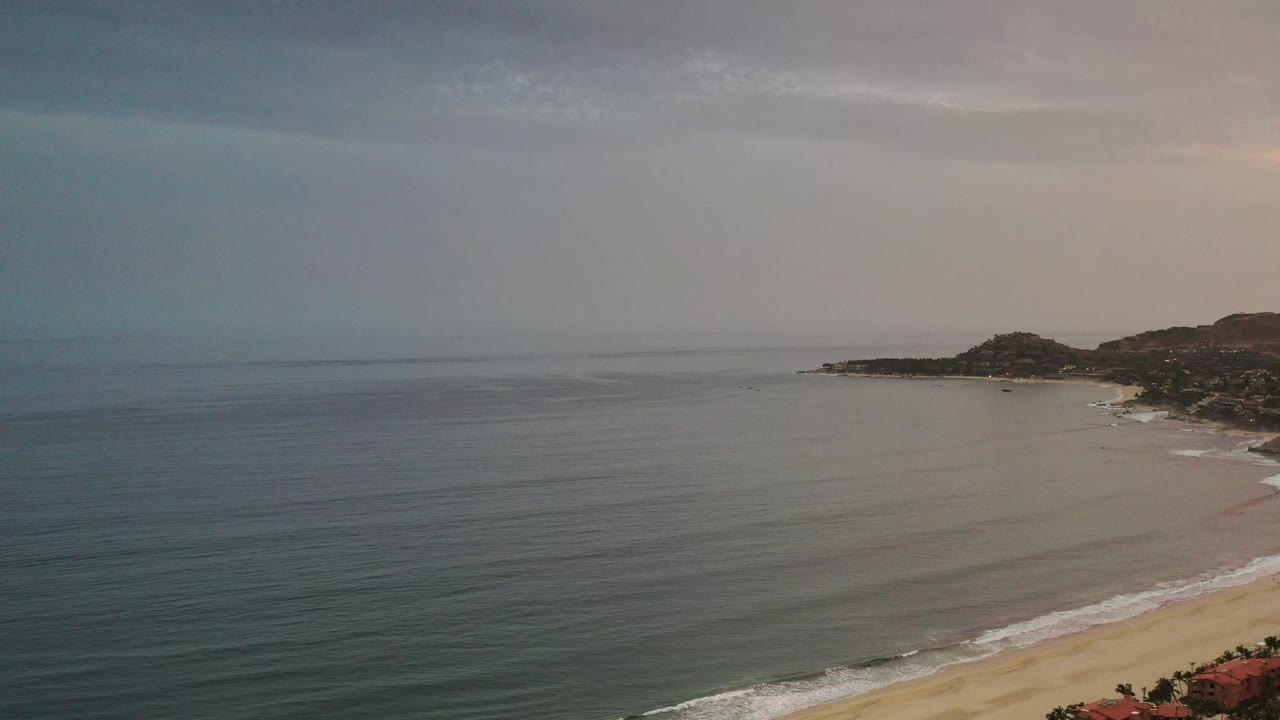 Los Cabos shoreline from above with calm ocean in the foreground, pale sandy beach curving along the right side, low hills beyond the inlet, and no visible people.