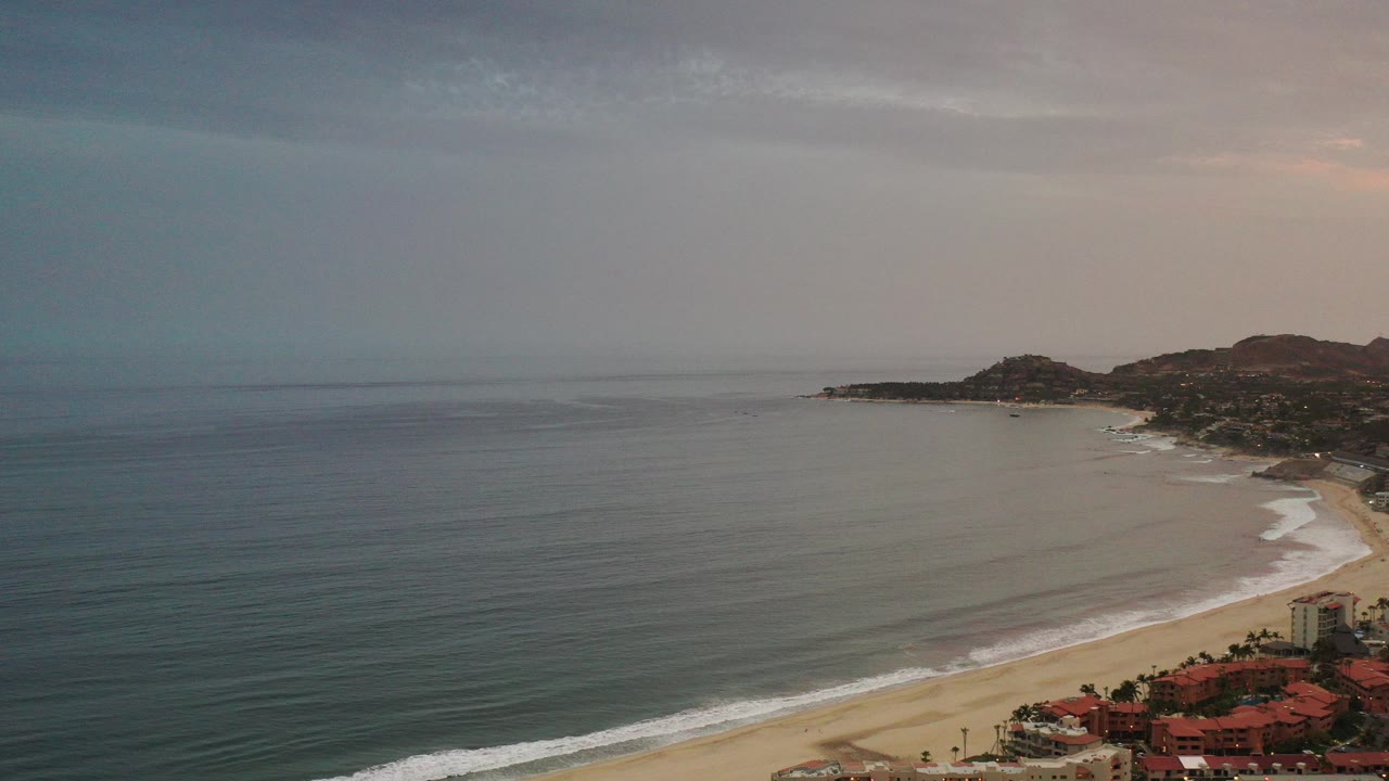 Aerial view of ocean, sandy beach, rooftops, and distant hills.