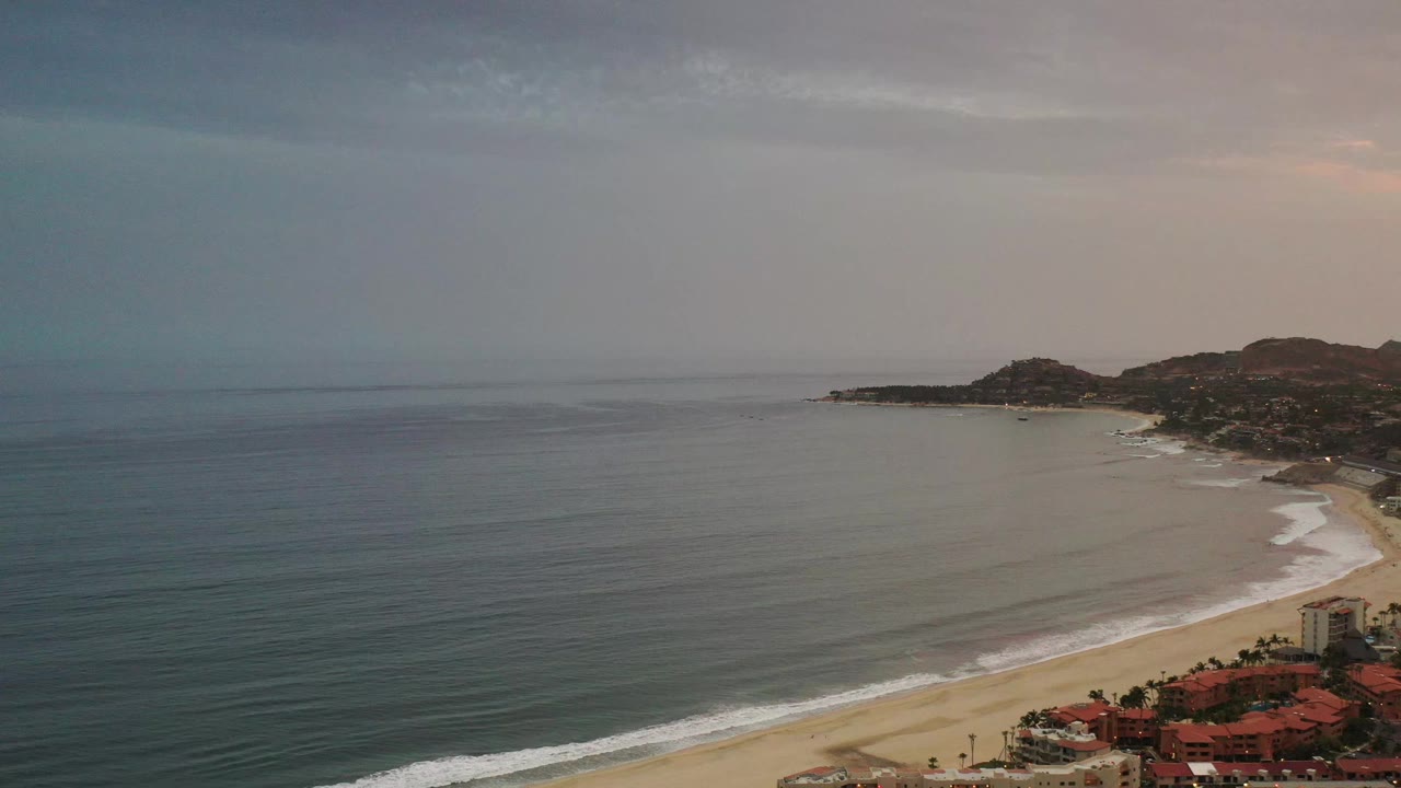 Early frame over Los Cabos, Mexico, with the calm ocean spread across the left, thin surf lines along the beach, clustered rooftops near the bottom edge, low hills beyond, and no visible people.