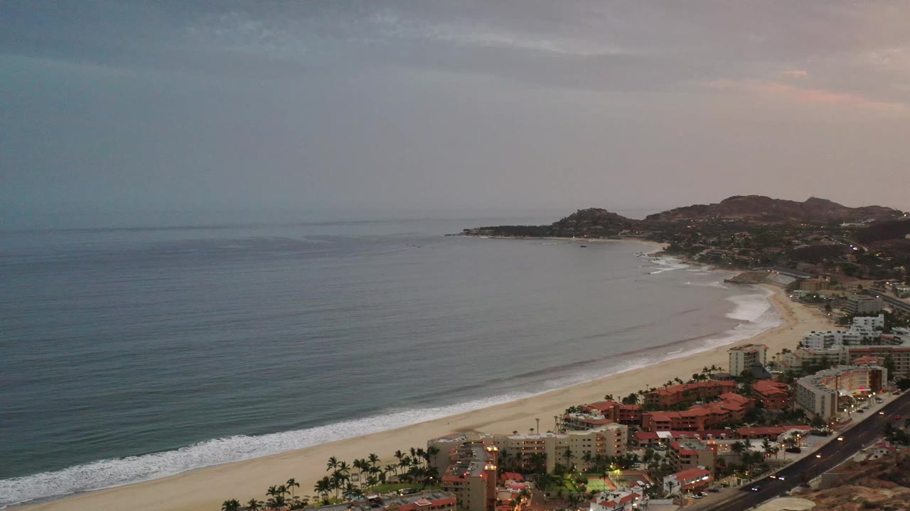 Aerial frame from Los Cabos, Mexico, showing the ocean stretching left, a pale beach curve, clustered beachfront buildings, hillside development, and no visible people.