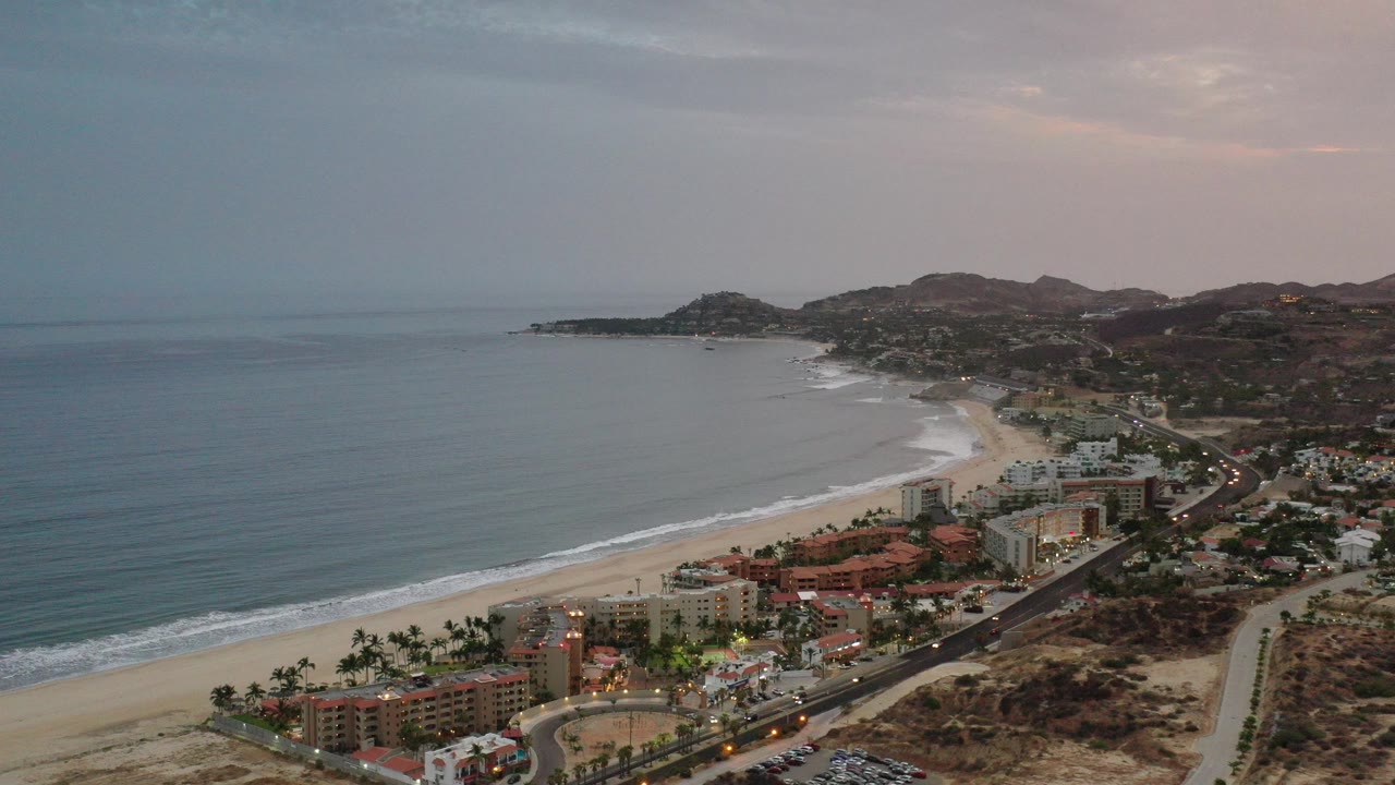 Aerial view of a curved beach, ocean, coastal buildings, and hills at dusk.