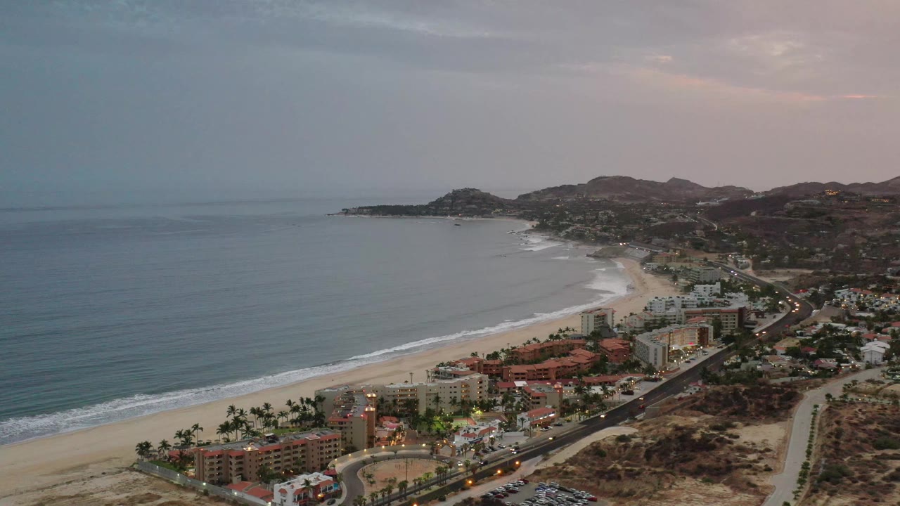 Aerial Los Cabos coastal frame with the ocean filling the left side, beach and palm trees below, hillside development to the right, and no visible people.