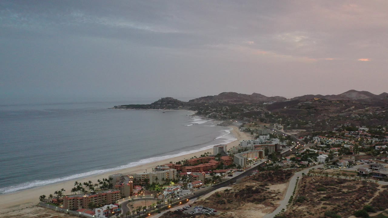 Aerial coastline with ocean, beach, buildings, roads, and hills at sunset.